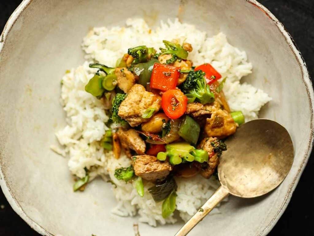 A white bowl filled with white rice topped with a colorful cashew nut stir fry including broccoli, red peppers, and pieces of tofu. A bronze spoon rests on the side of the bowl.